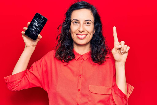 Joven mujer hispana hermosa sosteniendo teléfono inteligente roto mostrando pantalla quebrada sonriendo con una idea o pregunta apuntando con el dedo con la cara feliz, número uno.  - Foto, Imagen