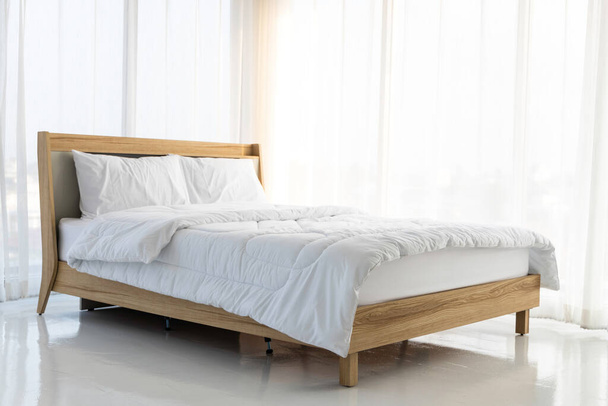 Το ξύλινο κρεβάτι είναι στρωμένο με λευκά σεντόνια σε άδειο δωμάτιο που περιβάλλεται από λευκές κουρτίνες και απαλό φως του ήλιου . - Φωτογραφία, εικόνα