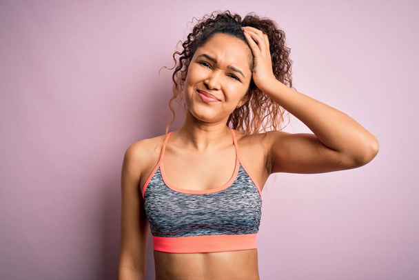 Beautiful sportswoman with curly hair doing sport wearing sportswear over pink background confuse and wonder about question. Uncertain with doubt, thinking with hand on head. Pensive concept. - Photo, Image