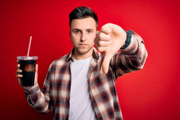 Fiatal jóképű kaukázusi férfi iszik egy friss üdítőt piros háttér dühös arc, negatív jel mutatja, nem tetszik hüvelykujjal lefelé, elutasítás koncepció - Fotó, kép