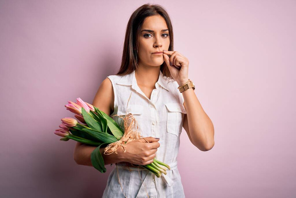 Jonge mooie brunette vrouw met boeket van tulpen bloemen over roze achtergrond mond en lippen gesloten als rits met vingers. Geheim en stil, taboe praten - Foto, afbeelding