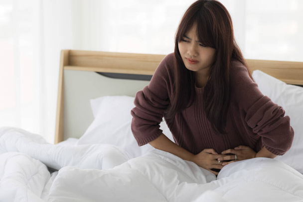 Asian woman is using her hands to hold her stomach because she has a stomach ache in bed room. Health care concept. - Foto, Imagem
