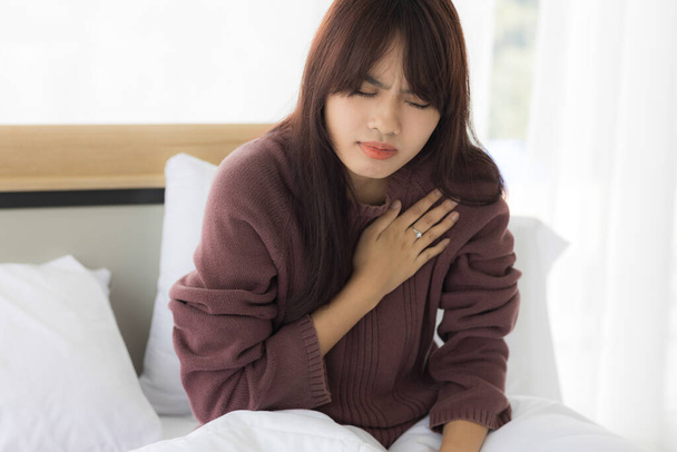 Yatak odasındaki Asyalı kadın ağırlaştırılmış kalp hastalığı nedeniyle kalbini tutmak için elini kullanıyor. Sağlık hizmeti kavramı. - Fotoğraf, Görsel