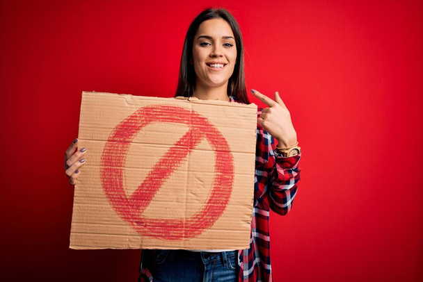 Young beautiful brunette woman holding banner with prohibited signal over red background with surprise face pointing finger to himself - Photo, image