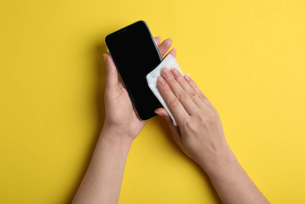 Woman cleaning mobile phone with antiseptic wipe on yellow background, top view - Foto, immagini
