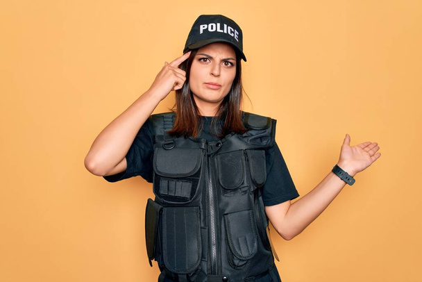 Jonge mooie brunette politievrouw draagt politie-uniform kogelvrij en pet verward en geïrriteerd met open palm tonen kopieerruimte en wijzende vinger naar het voorhoofd. Denk er eens over na.. - Foto, afbeelding