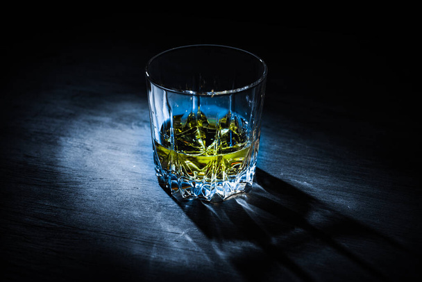 A glass of whiskey lit by a ray of light stands on a dark table and on a black background - Φωτογραφία, εικόνα