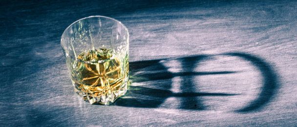 A ray of light illuminates a glass of whiskey standing on a blue wooden table, deep shadows from the sides of the glass, blue tone, copy space - 写真・画像
