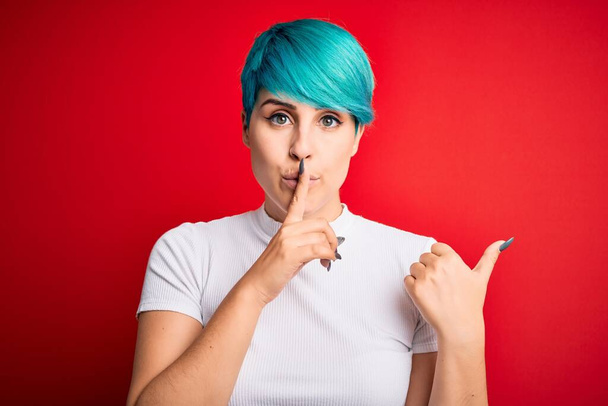 Junge schöne Frau mit blauem Modehaar in lässigem T-Shirt vor rotem Hintergrund, die darum bittet, mit dem Finger auf den Lippen, der mit der Hand zur Seite zeigt, ruhig zu sein. Schweigen und geheimes Konzept. - Foto, Bild