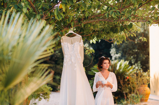 a beautiful bride stands next to a wedding dress with a Cup of tea in a boudoir outfit next to a Villa in Italy.morning of the bride in Tuscany. - Foto, afbeelding