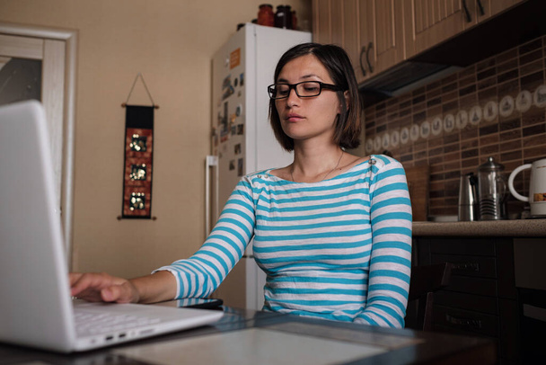 Νεαρή γυναίκα κάθεται στην κουζίνα και να παρακολουθήσουν webinar, να ακούσετε σε απευθείας σύνδεση πορεία, επικοινωνούν με τηλεδιάσκεψη κλήση βίντεο, e μάθουν τη γλώσσα στην εφαρμογή, μελέτη με μάθημα δάσκαλος ματιά στο φορητό υπολογιστή στο σπίτι - Φωτογραφία, εικόνα