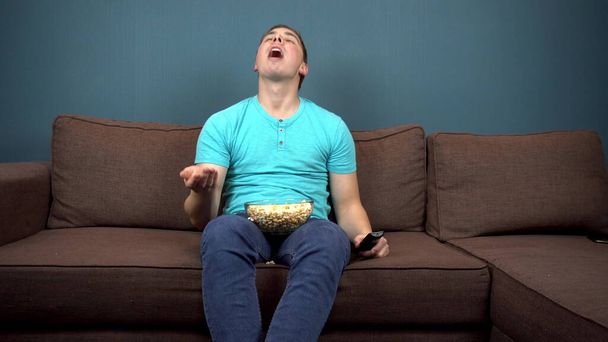 Een jongeman kijkt tv en eet popcorn. Hij vangt popcorn met z 'n mond. Zittend op de bank. Het uitzicht vanaf de TV. Kijkend naar de camera - Foto, afbeelding
