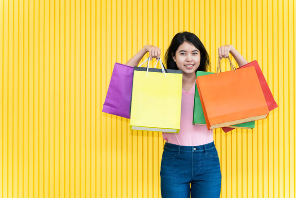 Asian young woman in a pink T-shirt and jeans is carrying several paper bags on yellow background. Shopping concept. - Foto, Imagem
