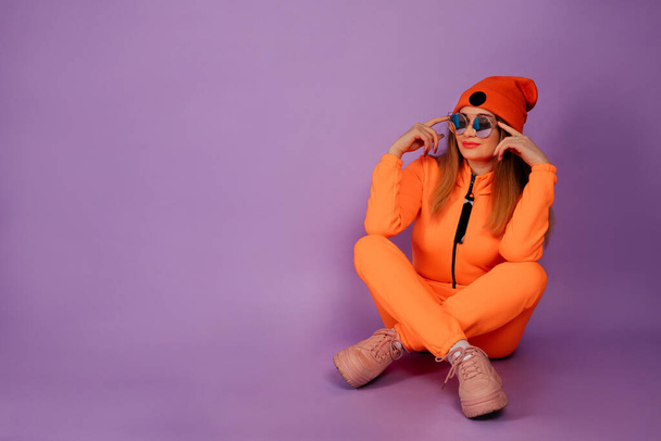 Mujer bastante joven sentada en elegante naranja en general, gorra y vidrio en aislado sobre fondo violeta. Foto de cuerpo entero
.  - Foto, Imagen