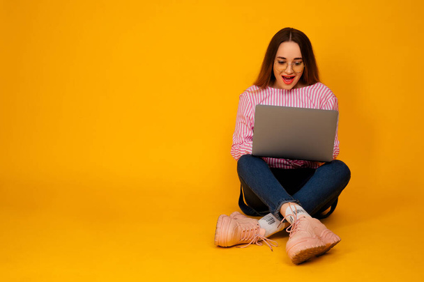 Ładna młoda kobieta na stylowej różowej koszulce, dżinsy z laptopem siedzącym na żółtym tle.  - Zdjęcie, obraz