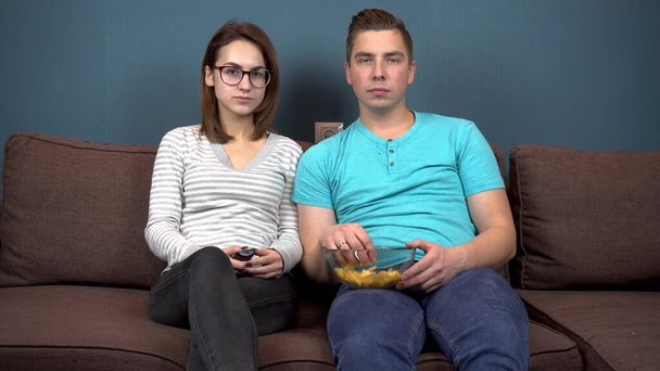 Мужчина и женщина смотрят телевизор и смеются. Молодая пара сидит на диване и ест чипсы. Стеклянная тарелка с чипсами в руках. Вид из телевизора. Смотреть в камеру
 - Фото, изображение