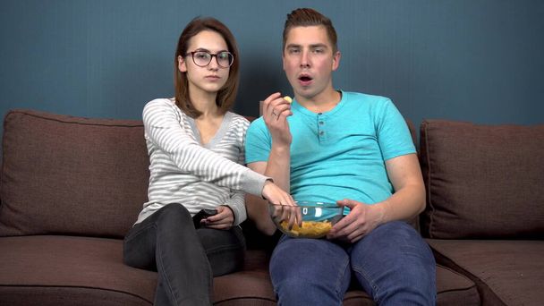 男と女がテレビを見て笑う。若いカップルがソファに座ってチップを食べています。手にチップとガラス板。テレビからの眺め。カメラを見ると - 写真・画像