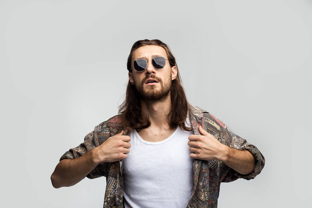 Brutal bully with long hair in dark sunglasses straightens his shirt, self-confident stylish guy on a white background, free style, people lifestyle - Photo, Image