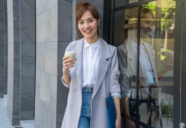 Güzel Asyalı kadın yürüyor, kahve içiyor, elinde bir fincan kahve tutuyor ve sabah işe gitmek için ofis binasının önünde duruyor.. - Fotoğraf, Görsel