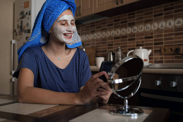 μια γυναίκα με μπλε πετσέτα στο κεφάλι μιλάει μέσω βίντεο σε ένα smartphone, πυροβολεί ένα vlog. μια λευκή πήλινη μάσκα στο πρόσωπό της. σπα στο σπίτι. ευεξία, πρωινό ξύπνημα και φροντίδα του σώματος έννοια. - Φωτογραφία, εικόνα
