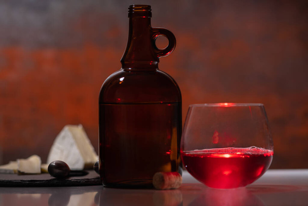 Glass of red liquor with bottle and cheese board highlighted by a beam of light on a bar counter in a low angle view with copy space - Фото, изображение