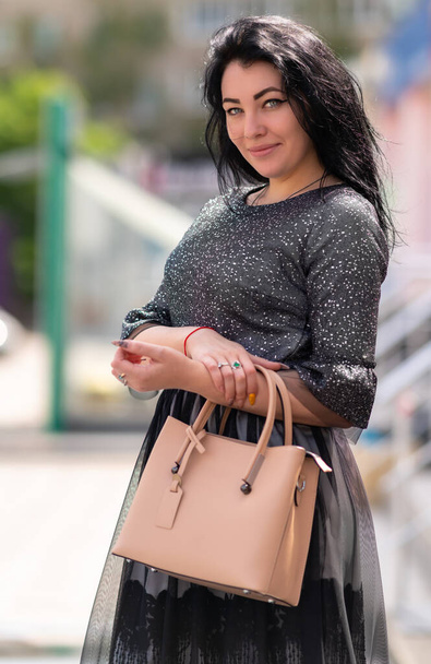 Smiling friendly attractive dark-haired woman posing with her handbag outdoors in town in the sunshine looking at the camera - Zdjęcie, obraz
