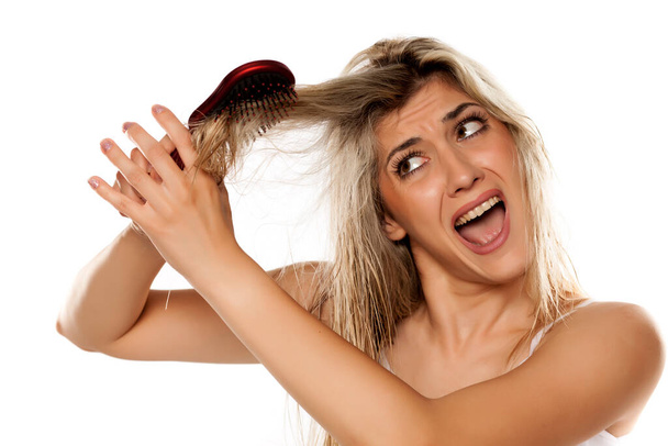 απελπισμένη ξανθιά γυναίκα χτενίζει τα βρώμικα βρεγμένα μαλλιά της σε λευκό φόντο - Φωτογραφία, εικόνα