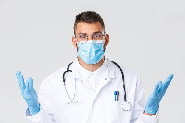 Covid-19, εργαζόμενοι στην υγειονομική περίθαλψη, πανδημία και πρόληψη της έννοιας του ιού. Κοντινό πλάνο ενός μπερδεμένου και σοκαρισμένου Ισπανόφωνου όμορφου γιατρού με γυαλιά, ιατρική μάσκα και γάντια, που δείχνει μπερδεμένος και ανίδεος. - Φωτογραφία, εικόνα