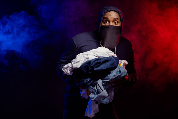 Portrait of scared frightened afro american guy rob boutique shop hold pile messy clothes horrified police chase him isolated over black fog color background - Fotó, kép