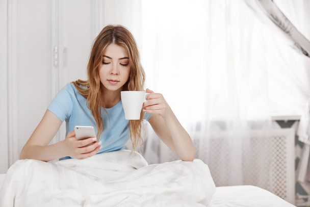 A beautiful young relaxed girl is sitting in bed in her pajamas. The girl drinks morning coffee and reads messages on her mobile phone - Foto, Imagen