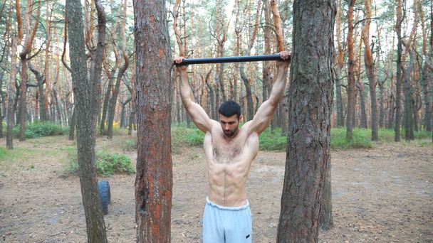 水平バーでいくつかの演習を示す強い男。筋肉質の男がプルアップしています。アスリートは森の中で筋力トレーニングを行う。スポーツマン訓練屋外。スポーツとアクティブライフスタイルの概念. - 写真・画像
