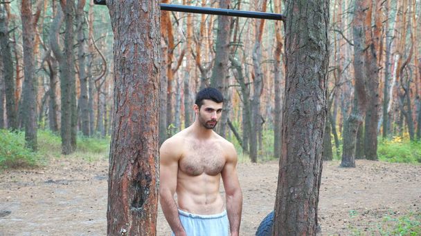 自然環境に立つ陸上競技マンの肖像画。筋肉の男のカメラを見て、水平バーでのトレーニングを開始します。アスリートは森の中で筋力トレーニングを行う。アクティブライフスタイルコンセプト. - 写真・画像