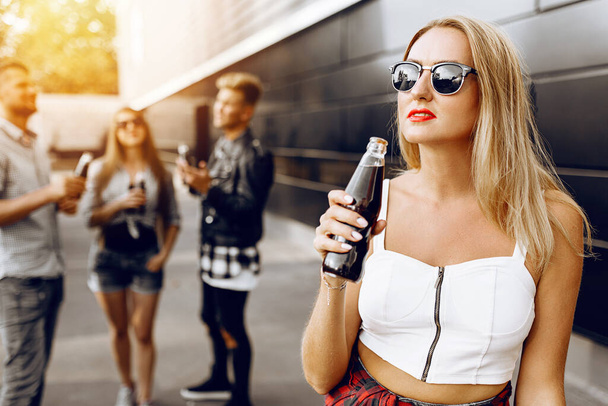 Close-up of a beautiful blonde girl with a glass drink, in the background a group of friends having fun, wearing sun glasses - Valokuva, kuva