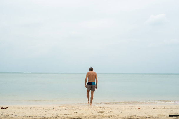 Young man with a long hair on a white sand beach and walks into tropical sea. Vacation and travel concept. - 写真・画像