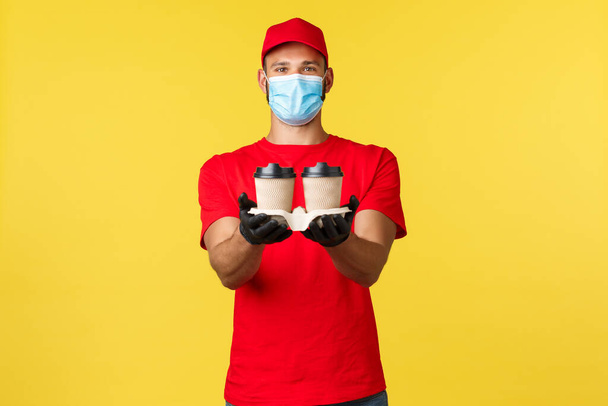 Takeaway delivery, food and drinks, covid-19 quarantine and preventing virus concept. Here your order. Friendly employee, courier in red uniform and medical mask, handing coffee to customer - 写真・画像