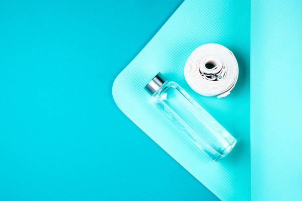 Top view of turquoise yoga mat, white belt and reusable glass water bottle on blue green background. Yoga pilates or fitness practice. Losing weight and sport concept. Flat Lay. Copy space. - Photo, Image