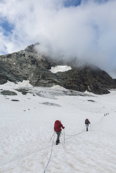 オーストリアの霧の中の岩の山に囲まれた雪のルート上のアルピニストグロスロックナー岩の頂上近くのアルプス, Kals am Grossglockner,オーストリア - 写真・画像