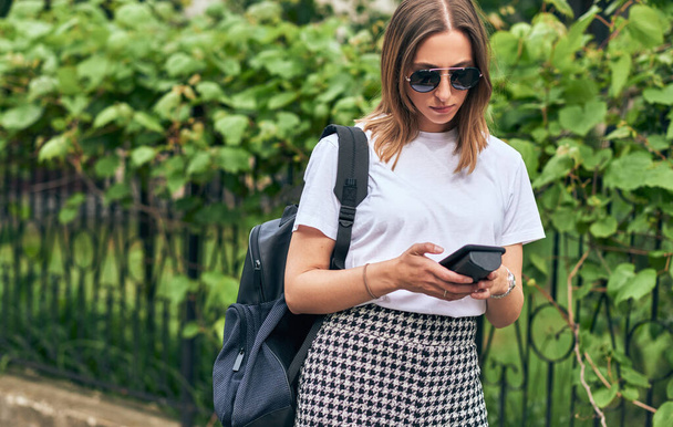Outdoor image of Caucasian young woman typing messages on smartphone, wearing white t-shirt, black sunglasses and backpack in the city street. Pretty female using free wireless on cellphone outside.  - Photo, Image