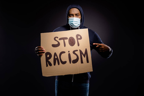 Photo of serious afro american guy hold point index finger card board banner share stop racism discrimination message wear jumper medical mask isolated black color background - Photo, image