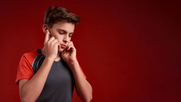 Concentration. A teenage boy engaged in sport, closing his eyes while listening to music via wireless earphones. Isolated on red background. Training, active lifestyle concept. Horizontal shot - Photo, image