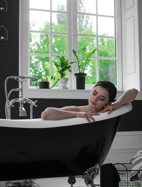 Uma linda jovem morena está em uma banheira branca contra uma parede de borgonha e uma janela branca com vasos de flores
 - Foto, Imagem