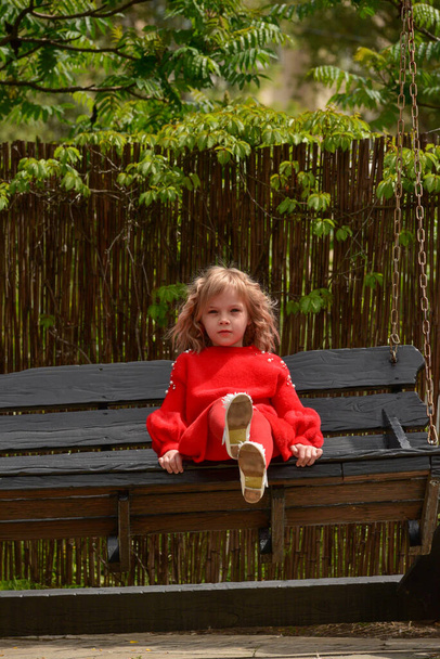 une blonde aux cheveux bouclés fille de 7 ans monte une grande balançoire en bois dans le parc. Fille sur une balançoire en bois. Une écolière en tenue rouge. Portrait d'enfant
. - Photo, image