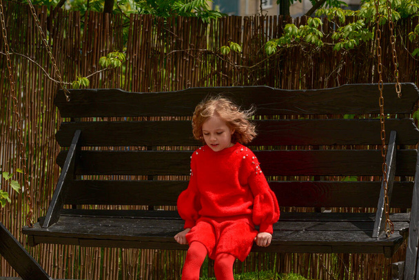 une blonde aux cheveux bouclés fille de 7 ans monte une grande balançoire en bois dans le parc. Fille sur une balançoire en bois. Une écolière en tenue rouge. Portrait d'enfant
. - Photo, image