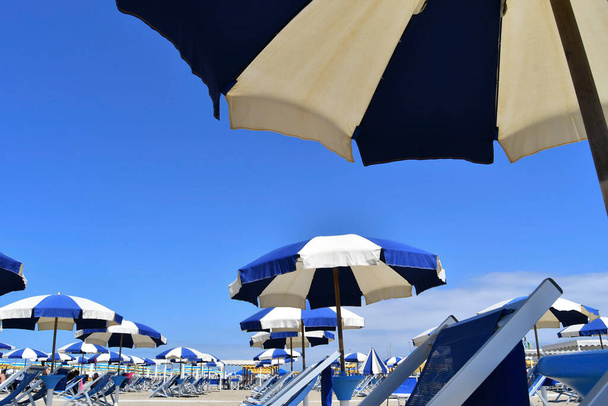 Avoin sateenvarjot ja hyvin vuorattu sininen ja valkoinen sbraes tuolit hiekkarannalla ilman ihmisiä
 - Valokuva, kuva