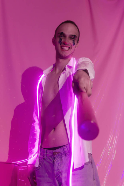 Egy fiatal jóképű, érzelmes férfi portréja, kigombolt fehér ingben, rózsaszín denevérrel. Férfi egy őrült bohóc képében, mint egy bohóc. Neon rózsaszín szoba. A neon tükörképe. Szelektív fókusz. - Fotó, kép
