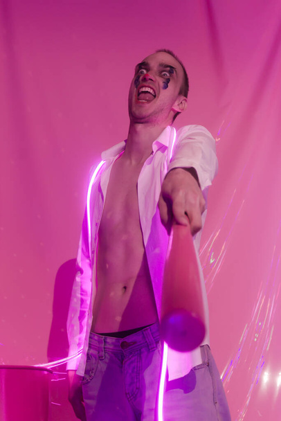 Portrait of Young Handsome emotional man in an unbuttoned white shirt with pink bat. Male in the image of a crazy clown like a joker. Neon pink room. Reflect of neon. Selective focus. - Photo, Image
