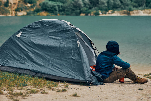 Young man sitting by the tent in nature and looking at beautiful lake. Weekend camping concept. - Photo, Image