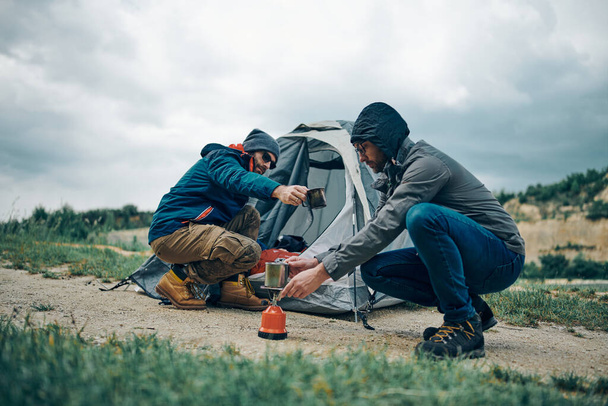 Двое привлекательных мужчин, приседающих у палатки на природе и готовящих утренний кофе
. - Фото, изображение