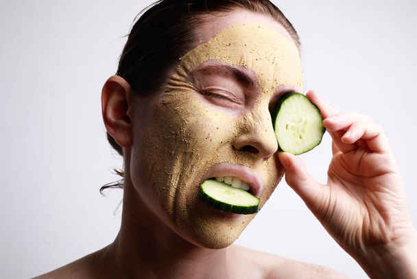 Woman with a face mask on holding a cucumber slices in front of one eye. Beauty and skincare concept. - Foto, imagen