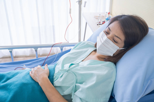 Azjatka jest chora ma wysoką gorączkę, kicha, dochodzi do siebie w sukience pacjenta leży na łóżku pacjenta w szpitalu z maskami na twarz chronić wirusa. koncepcja planowania19 - Zdjęcie, obraz